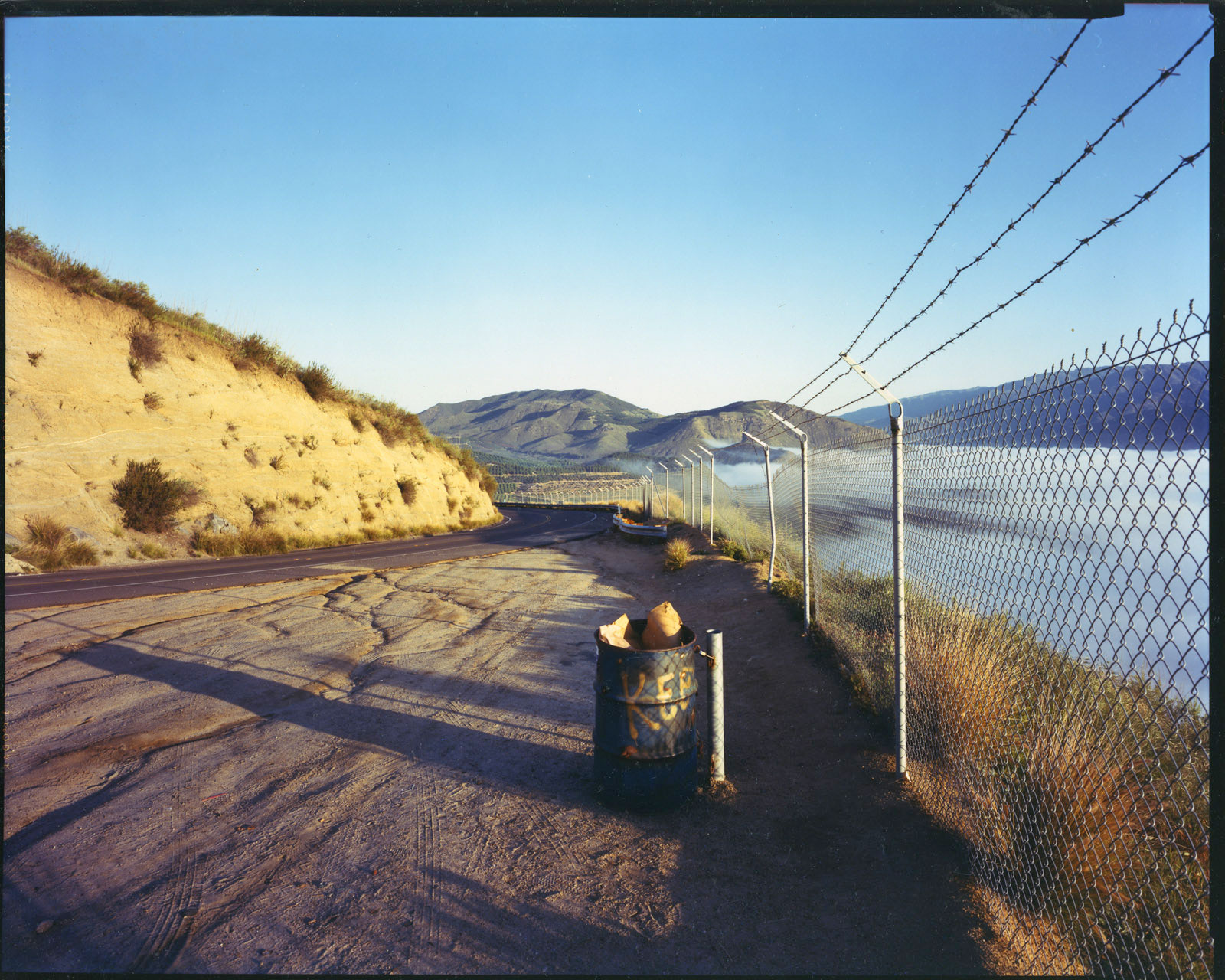 Hills in distance, eastern berm on left, chian-link fence on right, trash can center