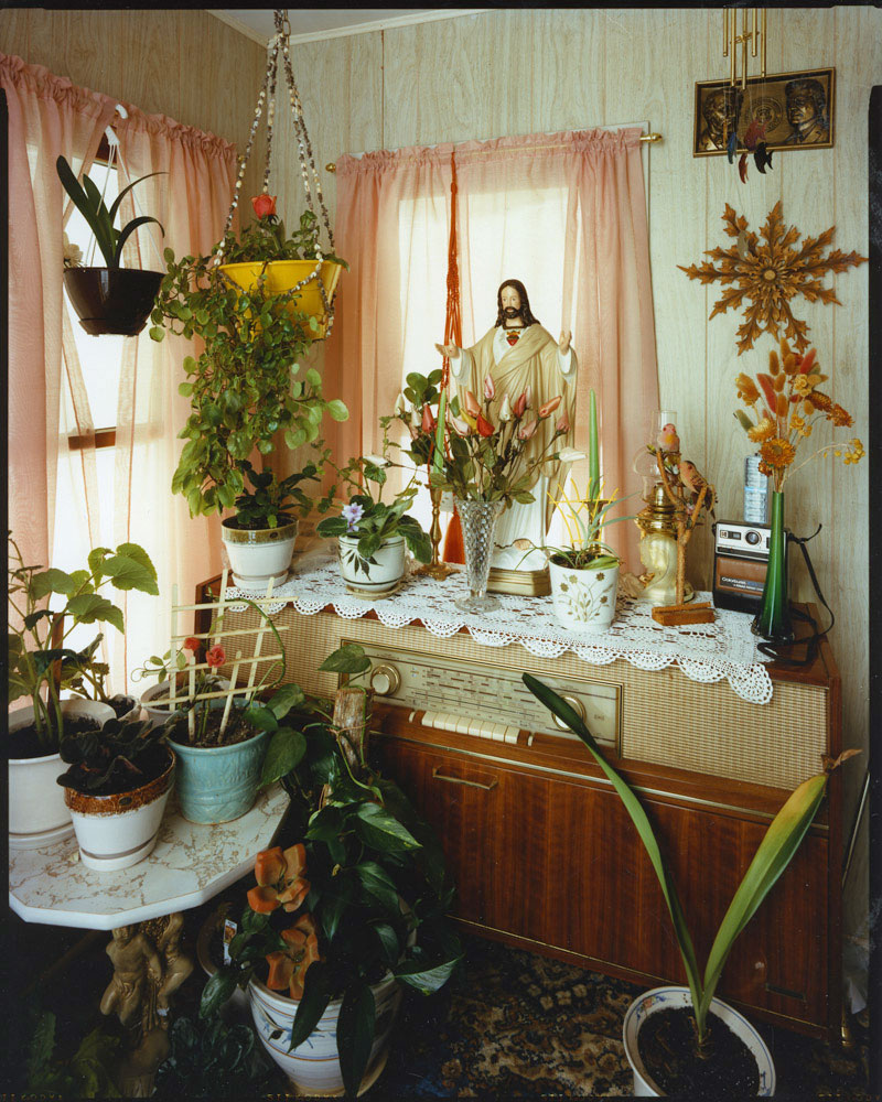 Jesus statue on top of stereo console, plants on console, table, and floor, in front of corner windows