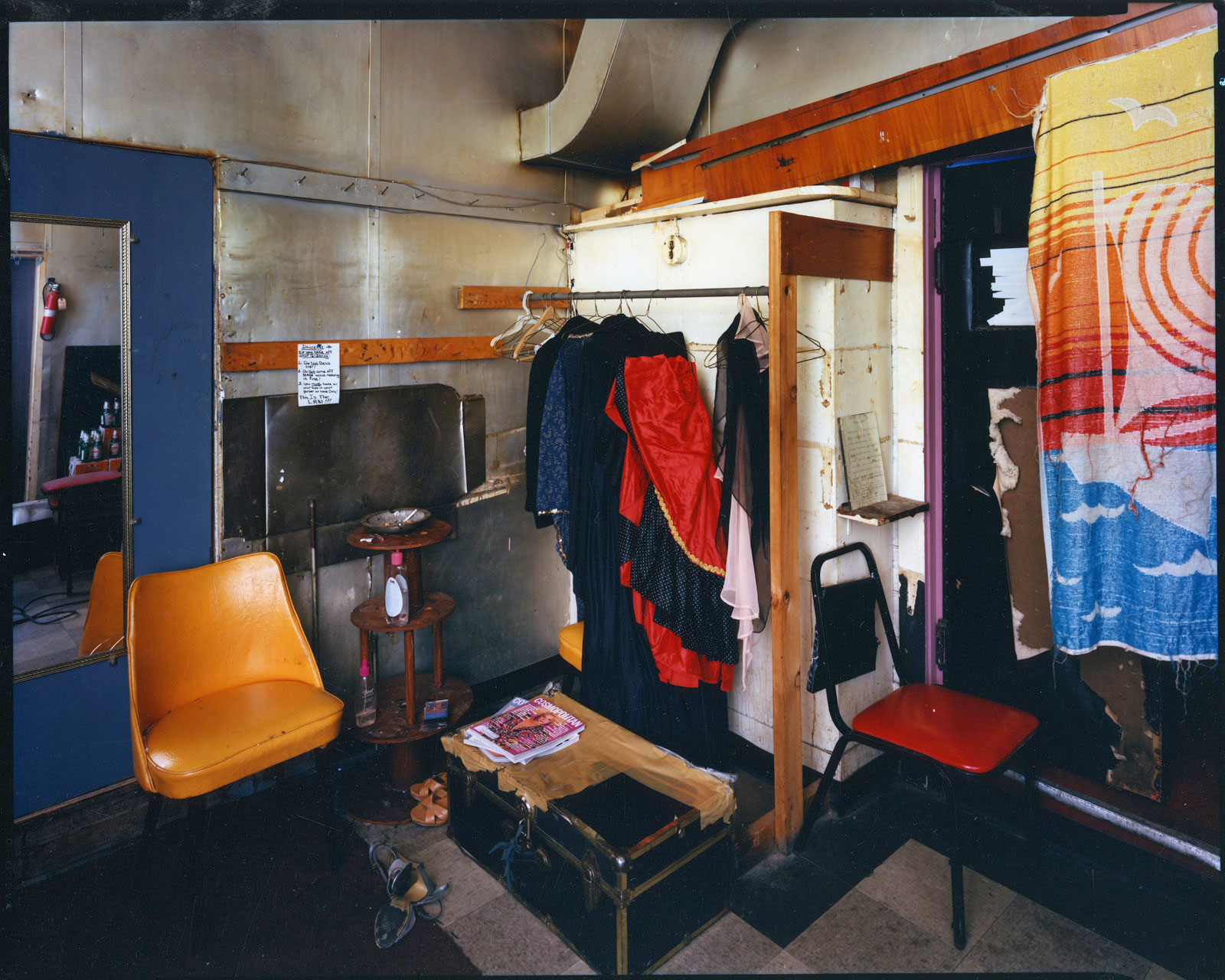 Gold chair on left, in front of mirror, trunk on floor, clothes hanging from rod