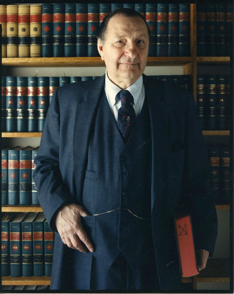 Man in 3-piece suit, holding book, in front of library shelves