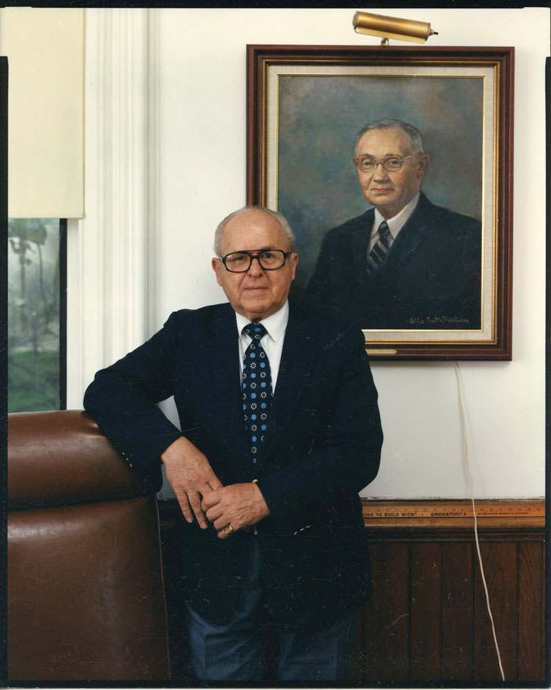 Man in dark suit in front of framed portrait on wall