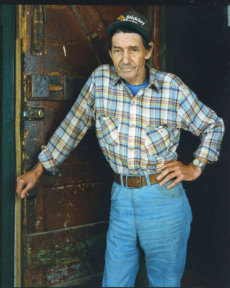 Man wearing plaid shirt, blue jeans, and Strickley cap, right hand leaning on doorknob