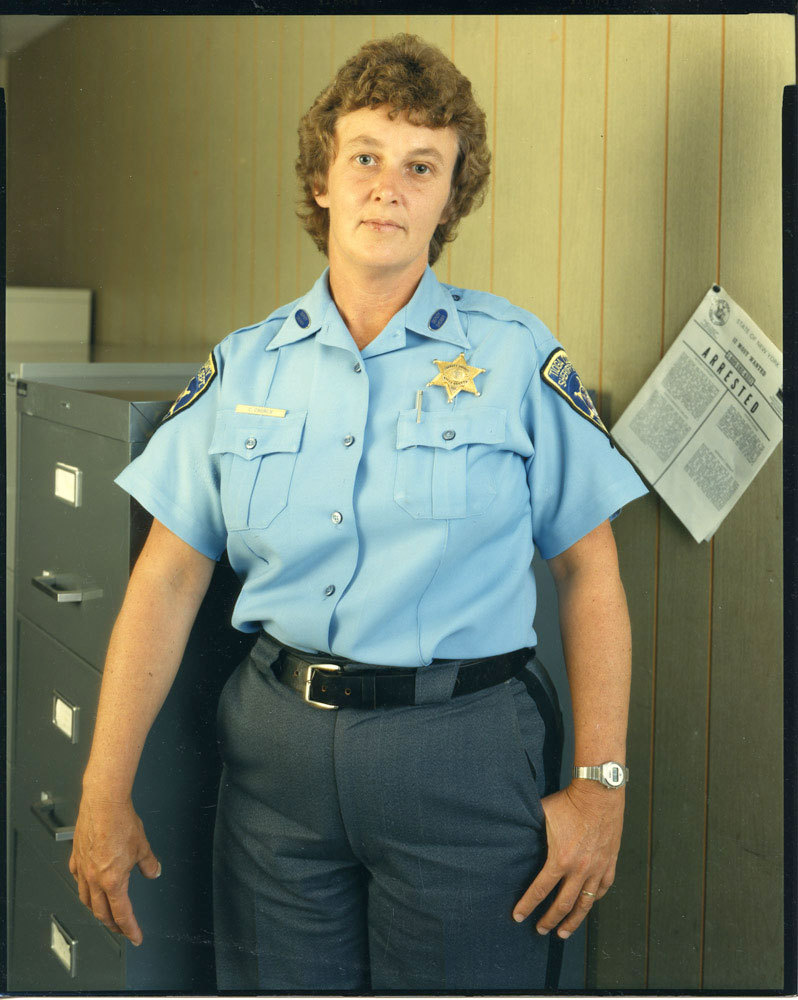 Woman in blue Tioga County Sheriff uniform, with file cabinet on left and ARRESTED noticed on right